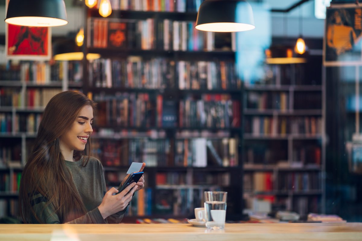 Busy woman sitting in a cafe with credit card and smartphone while while paying bills online.Woman pays for purchases with a card and phone. Makes shopping in popular web store.